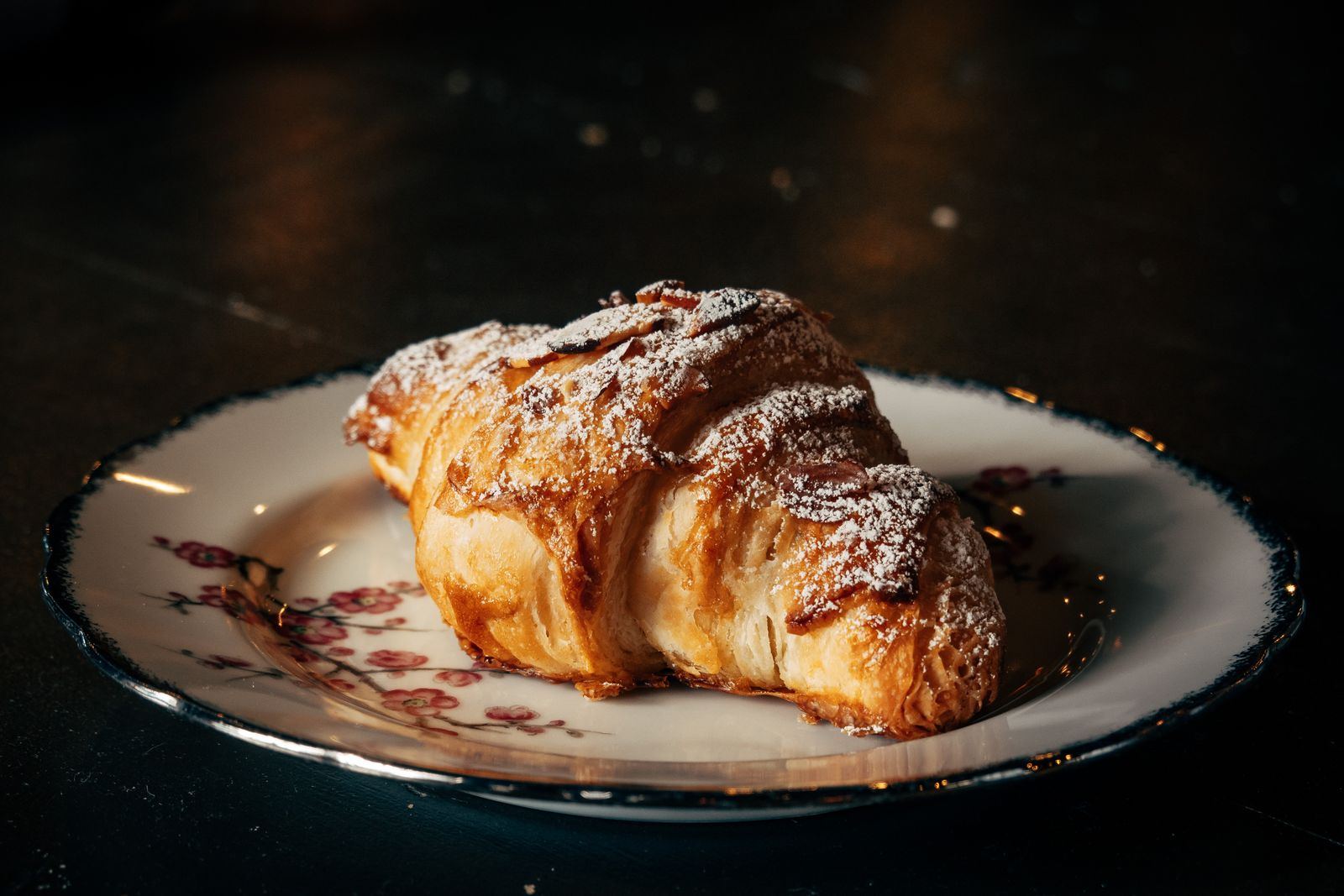 An almond croissant dusted with sugar on a plate, on a dark counter